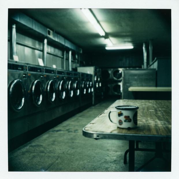 Polaroid of a laundromat interior at night, a row of front-loading machines and a chipped enamel mug on the table